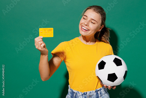 Young happy woman fan in basic yellow t-shirt cheer up support football sport team hold soccer ball yellow card propose player retire from field watch tv live stream isolated on dark green background. photo