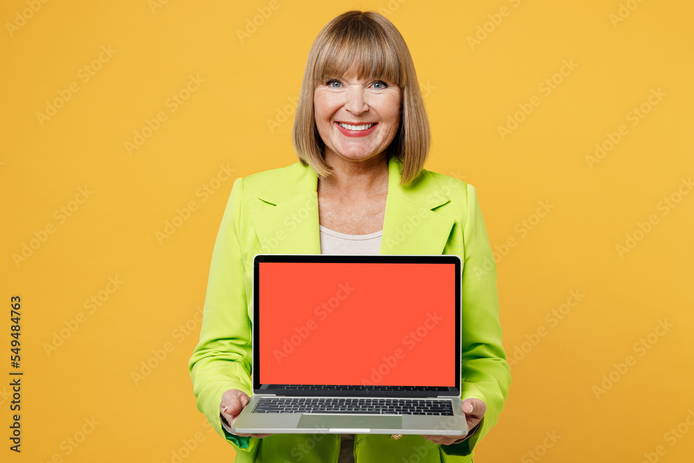Naklejka premium Elderly smiling happy IT woman 50s years old wear green jacket white t-shirt hold use work on laptop pc computer with blank screen workspace area isolated on plain yellow background studio portrait.
