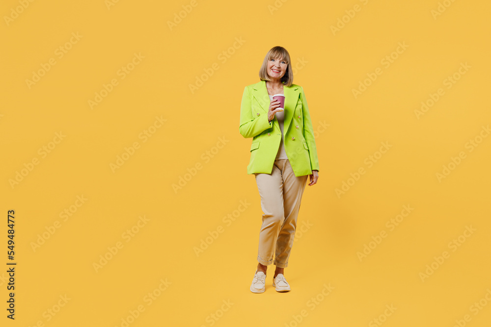 Full body elderly woman 50s years old wear green jacket white t-shirt hold takeaway delivery craft paper cup coffee to go isolated on plain yellow background studio portrait. People lifestyle concept. Full body elderly woman 50s years old wear green jacket white t-shirt hold takeaway delivery craft paper cup coffee to go isolated on plain yellow background studio portrait. People lifestyle concept.