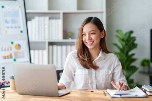 Pretty and cute Asian businesswoman smiling and enjoying coming up with ideas for working with laptop and document. © amnaj