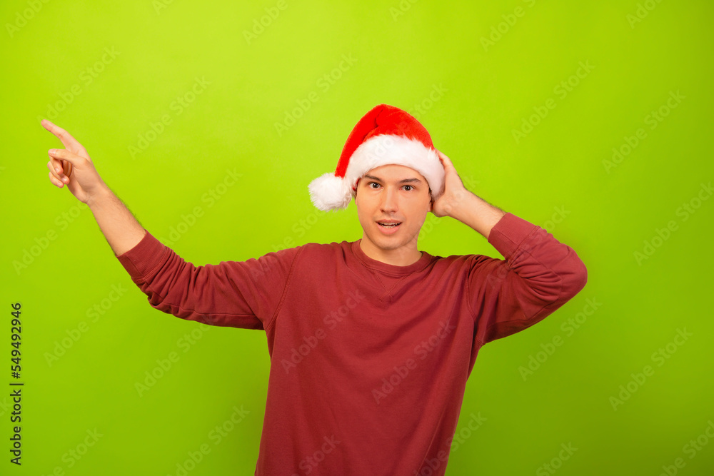 Surprised amazed young man wearing Santa Claus hat, red casual sweater, demonstrate or showing offer ads promo, pointing aside with fingers hand gesture and looking at camera on green background.