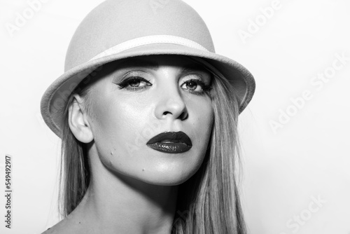 Fashion and make-up concept. Studio portrait of beautiful blonde woman with efancy make-up. Model wearing retro hat and looking at camera with serious and seductive look. black and white image