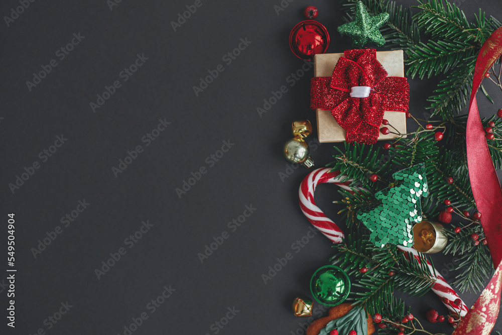 Stylish christmas gift, tree branches, candy cane, festive decorations border on black background. Modern christmas flat lay, space for text. Season's greetings card. Merry Christmas!