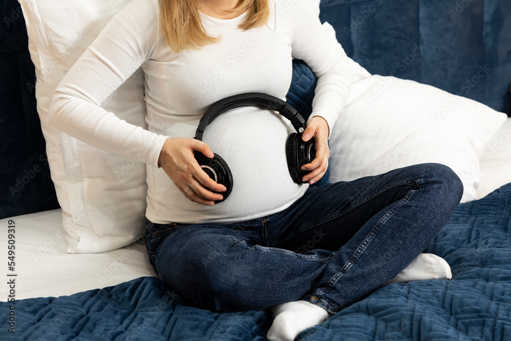 Young pretty pregnant woman holds black headphones on belly sitting on bed,  prenatal music listening. Happy childbearing period, caring for unborn  baby. Female wears white long sleeve, blue jeans Stock Photo