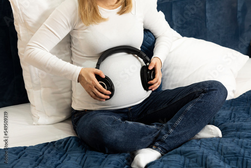 Young pretty pregnant woman holds black headphones on belly sitting on bed, prenatal music listening. Happy childbearing period, caring for unborn baby. Female wears white long sleeve, blue jeans photo