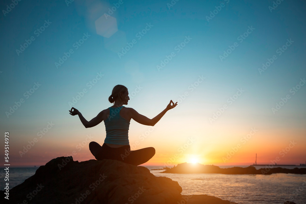 A woman doing yoga in the lotus pose meditating on the beach during sunset.