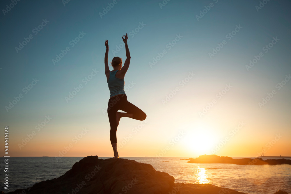 A woman doing yoga, meditating on the beach during sunset.