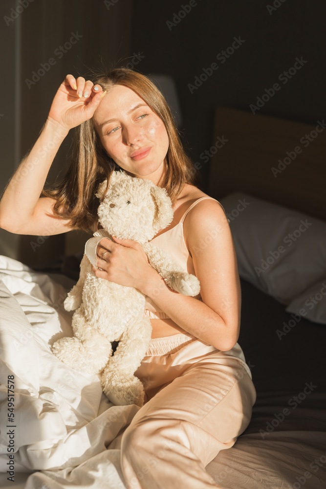 Girl is waking up in morning stretches in bed and sun shines from window. Happy young woman greets new sunny day and holds teddy bear toy Girl is waking up in morning stretches in bed and sun shines from window. Happy young woman greets new sunny day and holds teddy bear toy