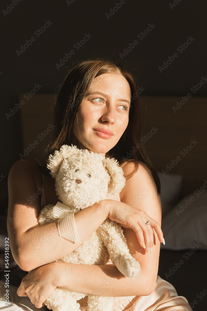 Portrait of girl is waking up in morning stretches in bed and sun shines from window. Happy young woman greets new sunny day and holds teddy bear toy Portrait of girl is waking up in morning stretches in bed and sun shines from window. Happy young woman greets new sunny day and holds teddy bear toy