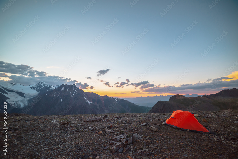 Scenic alpine landscape with tent at very high altitude with view to large mountains in orange dawn sky. Vivid orange tent with awesome view to high mountain range under cloudy sky in sunset colors.