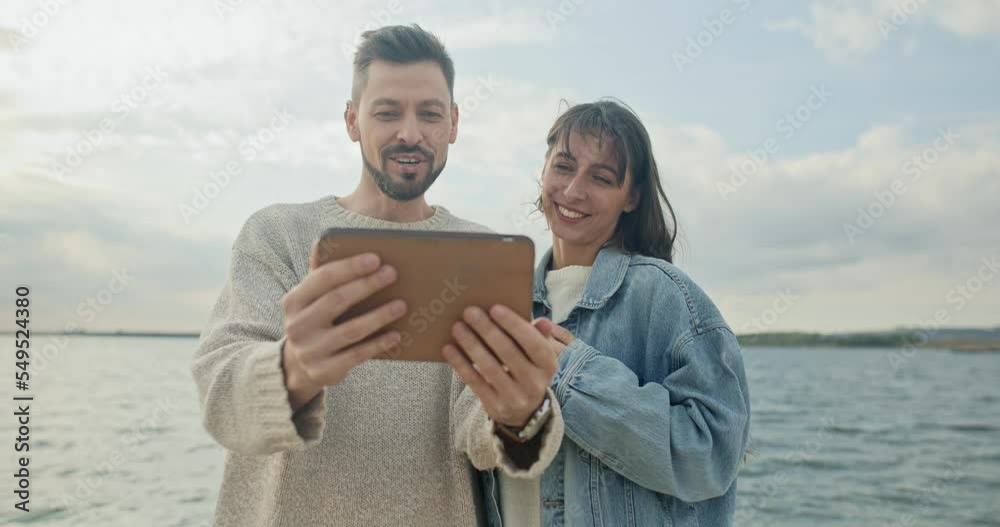 Young cute woman and beautiful elegant man using tablet video calling talking online standing on beach near a beautiful big lake enjoying trip. Lovely happy couple communicating smiling. Young cute woman and beautiful elegant man using tablet video calling talking online standing on beach near a beautiful big lake enjoying trip. Lovely happy couple communicating smiling.