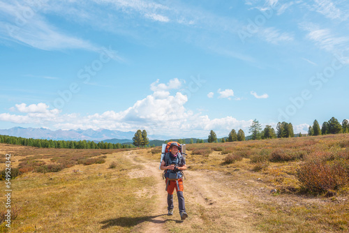 Alone traveler with large backpack walks along hiking trail on sunlit high mountain plateau under white clouds in blue sky. Backpacker with photo camera in autumn mountain trekking in good weather. © Daniil
