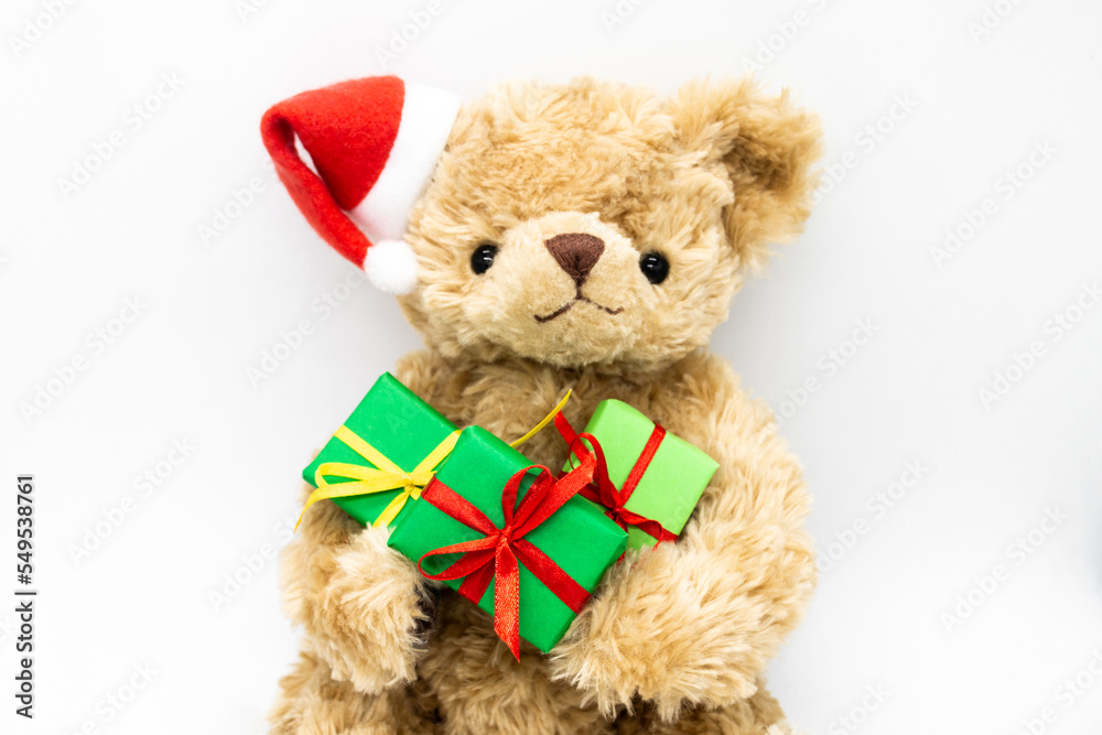 A stuffed toy Teddy bear in a red Santa Claus hat with a pompom on one ear, holding green gift boxes in its paws. White background, copy space.