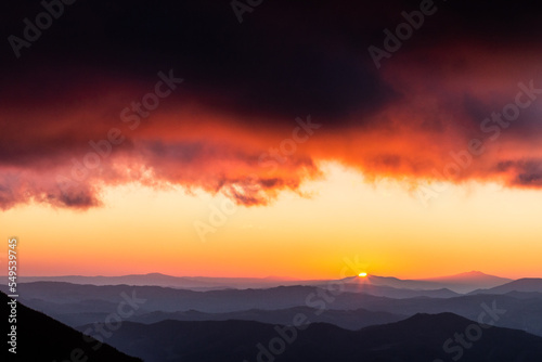 Distant sunset above layers of mountains and valleys with mist and fog, with a rooftop of close red clouds