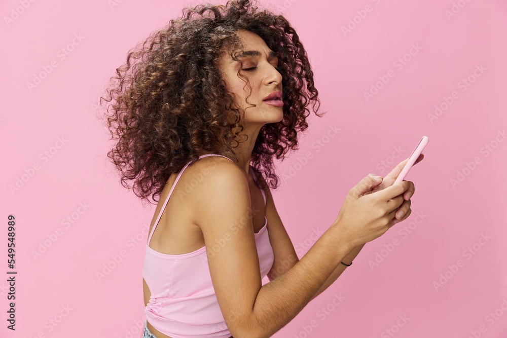 Woman blogger holding phone with curly hair in pink top and jeans poses on pink background, copy space, technology and social media Woman blogger holding phone with curly hair in pink top and jeans poses on pink background, copy space, technology and social media
