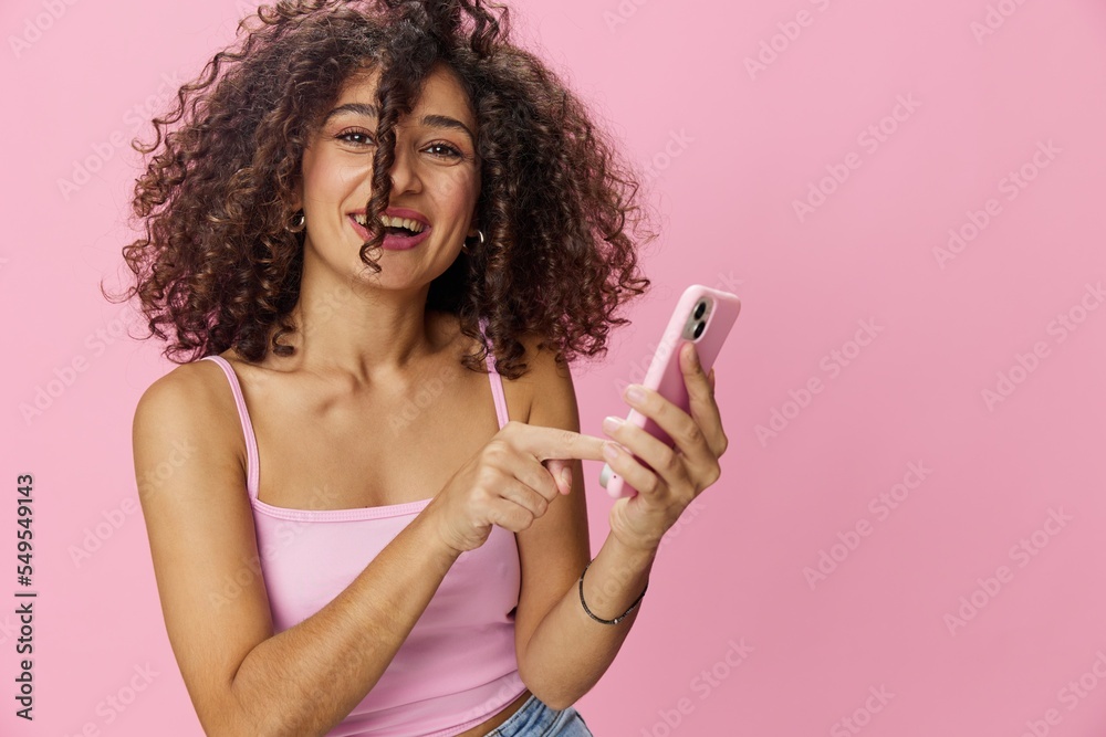 Woman blogger holding phone in hand video call, with curly hair in pink top and jeans poses on pink background, copy space, technology and social media, online Woman blogger holding phone in hand video call, with curly hair in pink top and jeans poses on pink background, copy space, technology and social media, online