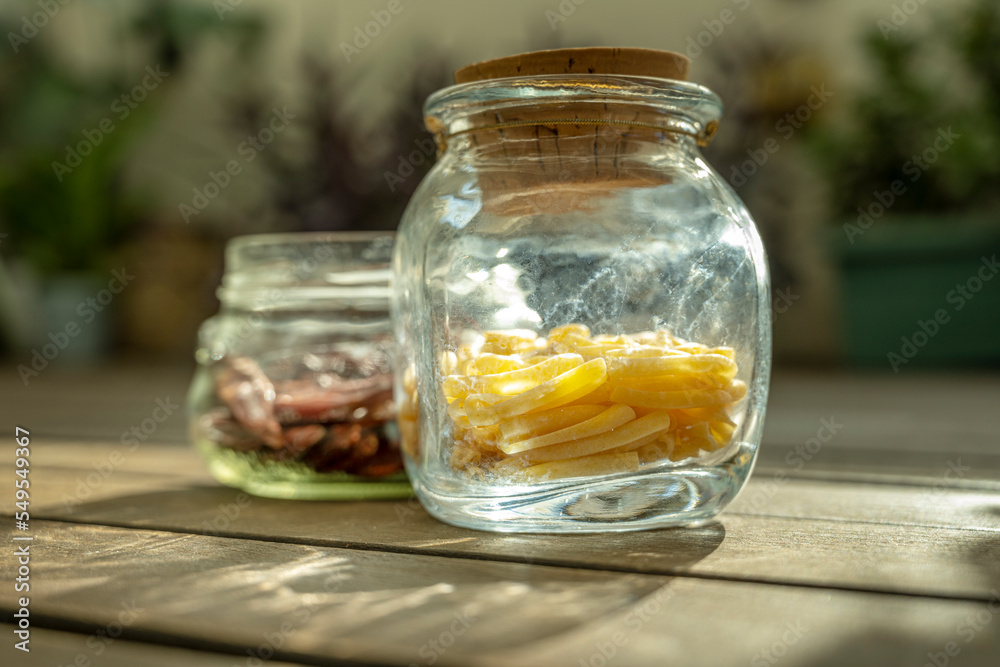 Some glass jars full of pasta with a cork stopper and another without a stopper and with dried chili peppers illuminated by a ray of morning sun Some glass jars full of pasta with a cork stopper and another without a stopper and with dried chili peppers illuminated by a ray of morning sun