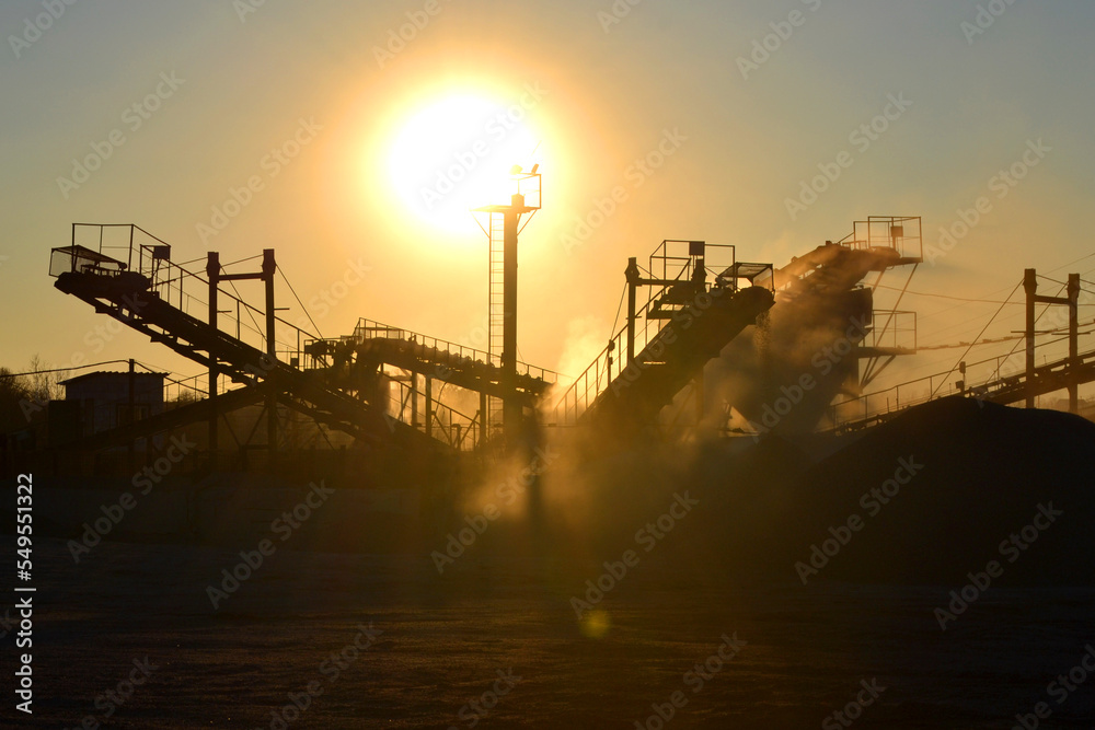 Mining quarry. Crushing plant in granite quarry at sunset dawn. Crusher in quarry. Lot of dust. Stone crushing. Industrial production of gravel. Gravel conveyor. Cone type rock crusher, shining sun