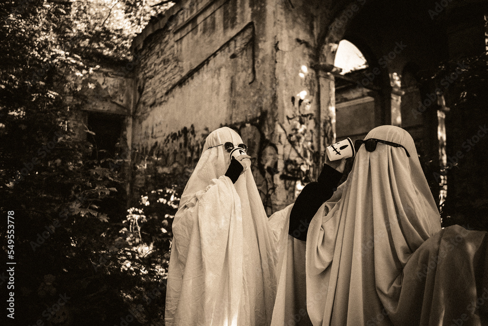 A funny image of two people in ghost costumes and sunglasses holding a cup with a drink in an abandoned building, black and white image