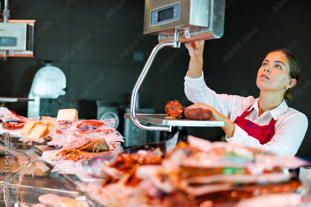 Positive female seller weighing sausage on scale in butcher shop