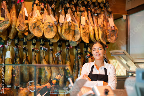 Portrait of positive young female butcher on background of rack with hanging various jamon in shop
