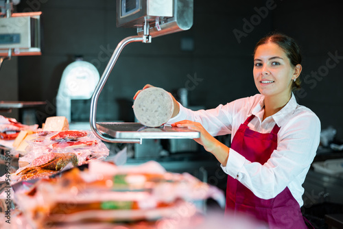 Positive young female seller of local grocery store standing behind counter, weighing traditional spanish mortadella on scales photo