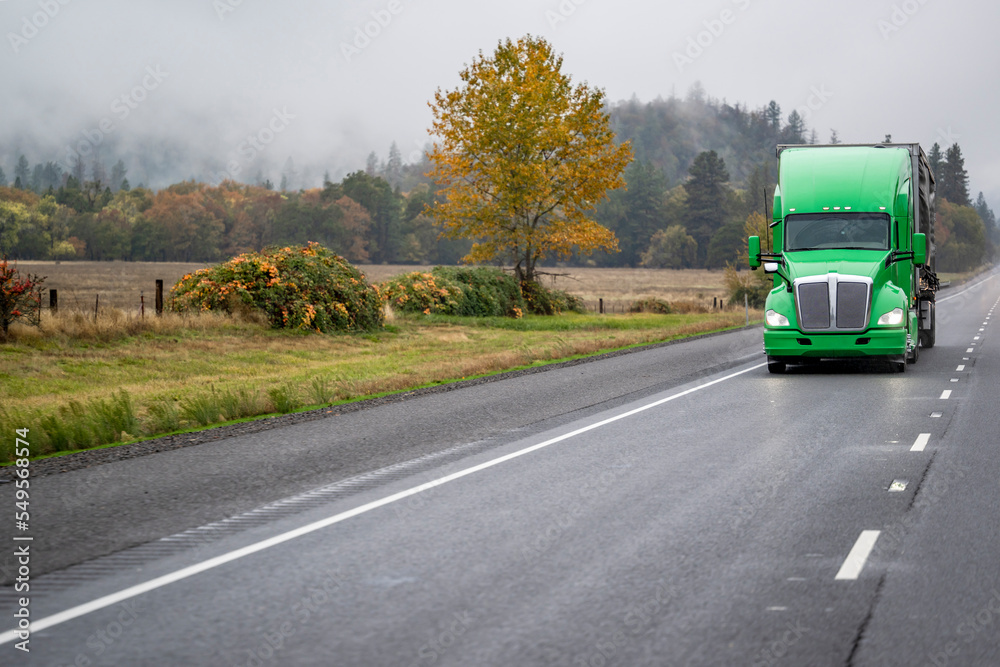 Green wet big rig bonnet semi truck transporting cargo in covered semi trailer driving on the slippery highway road at heavy raining weather Green wet big rig bonnet semi truck transporting cargo in covered semi trailer driving on the slippery highway road at heavy raining weather