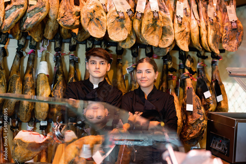 Two confident positive sellers of butcher shop, young girl and guy wearing black uniform, standing with crossed arms behind counter near rack with hanging whole legs of dry-cured Iberian jamon photo