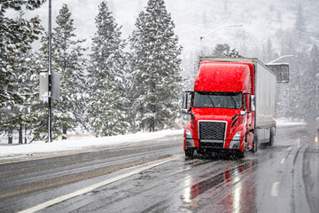 Industrial bonnet bright red big rig semi truck with dry van loaded semi trailer slipping slowly and carefully on a snowy winter highway near Shasta Lake in California