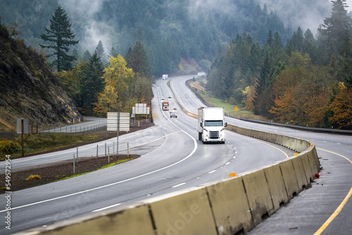 Industrial big rig semi truck with semi trailer transporting cargo driving on the winding highway road with fog and forest