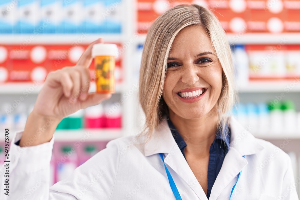 Young blonde woman pharmacist smiling confident holding pills bottle at pharmacy