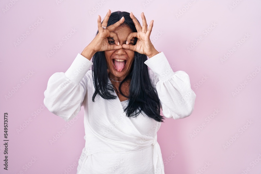 Mature hispanic woman standing over pink background doing ok gesture like binoculars sticking tongue out, eyes looking through fingers. crazy expression. Mature hispanic woman standing over pink background doing ok gesture like binoculars sticking tongue out, eyes looking through fingers. crazy expression.