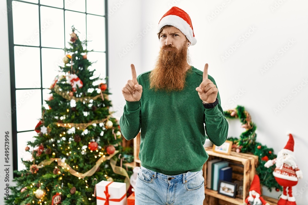 Redhead man with long beard wearing christmas hat by christmas tree pointing up looking sad and upset, indicating direction with fingers, unhappy and depressed. Redhead man with long beard wearing christmas hat by christmas tree pointing up looking sad and upset, indicating direction with fingers, unhappy and depressed.