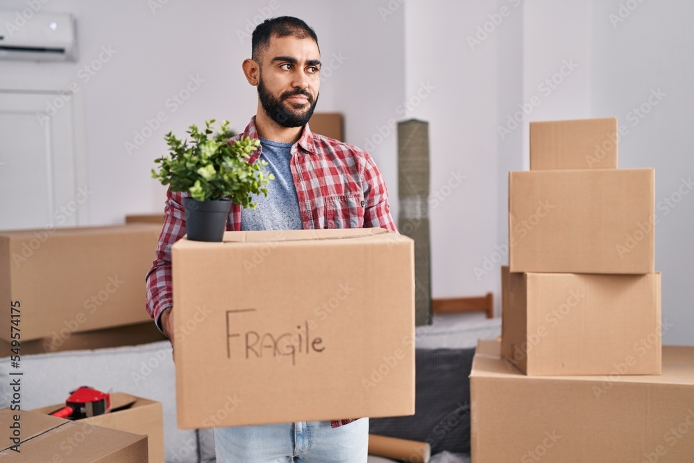 Middle east man with beard moving to a new home holding cardboard box smiling looking to the side and staring away thinking.