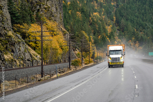 White big rig industrial long haul semi truck transporting oversize load driving on the autumn road with rock mountain at raining weather