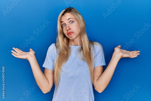 Beautiful blonde woman wearing casual t shirt over blue background clueless and confused expression with arms and hands raised. doubt concept.