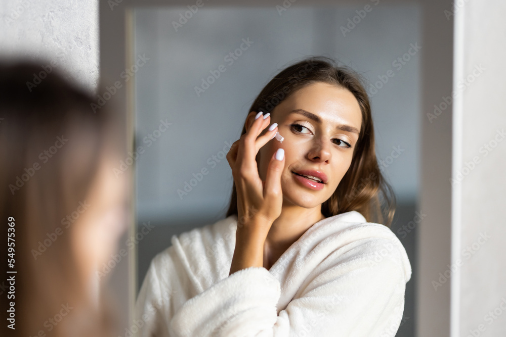 Close up cropped portrait of a young beautiful woman in bath towel smiling applying face body cream on her face for rejuvenation soft moisturizing effect. Spa beauty treatment concept. Body skin care Close up cropped portrait of a young beautiful woman in bath towel smiling applying face body cream on her face for rejuvenation soft moisturizing effect. Spa beauty treatment concept. Body skin care