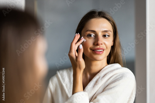Close up cropped portrait of a young beautiful woman in bath towel smiling applying face body cream on her face for rejuvenation soft moisturizing effect. Spa beauty treatment concept. Body skin care