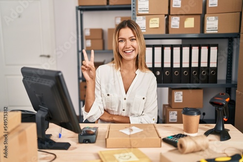 Young blonde woman working at small business ecommerce smiling with happy face winking at the camera doing victory sign. number two.