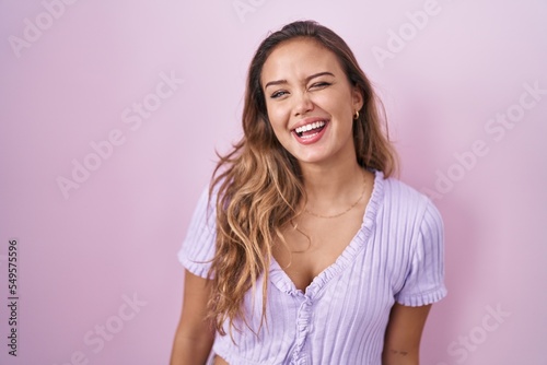 Young hispanic woman standing over pink background winking looking at the camera with sexy expression  cheerful and happy face.