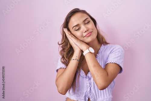 Young hispanic woman standing over pink background sleeping tired dreaming and posing with hands together while smiling with closed eyes.