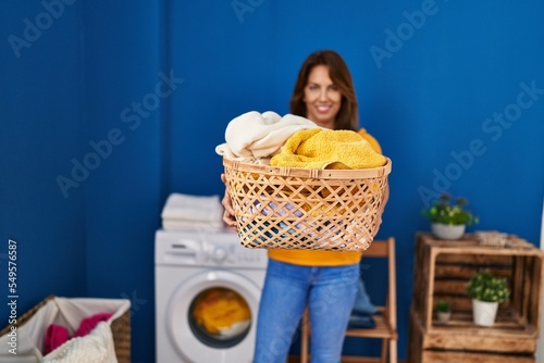 Young latin woman smiling confident holding basket with clothes at laundry room
