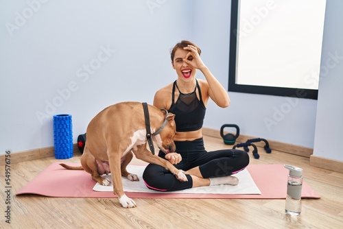 Young beautiful woman sitting on yoga mat doing ok gesture with hand smiling, eye looking through fingers with happy face.