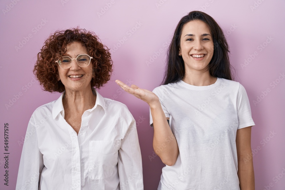 Hispanic mother and daughter wearing casual white t shirt over pink background smiling cheerful presenting and pointing with palm of hand looking at the camera. Hispanic mother and daughter wearing casual white t shirt over pink background smiling cheerful presenting and pointing with palm of hand looking at the camera.