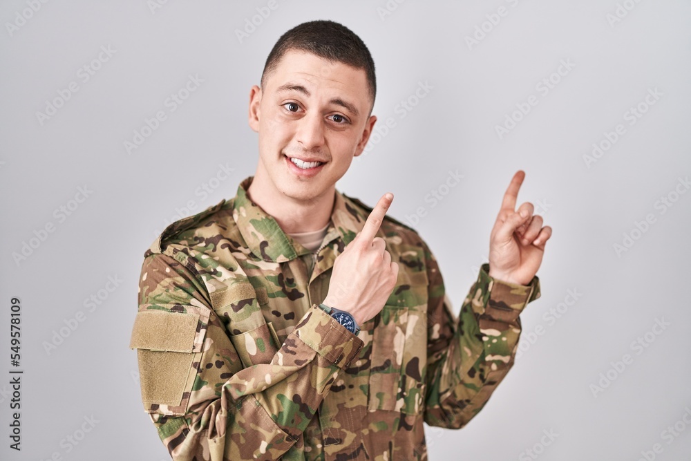 Young man wearing camouflage army uniform smiling and looking at the camera pointing with two hands and fingers to the side.