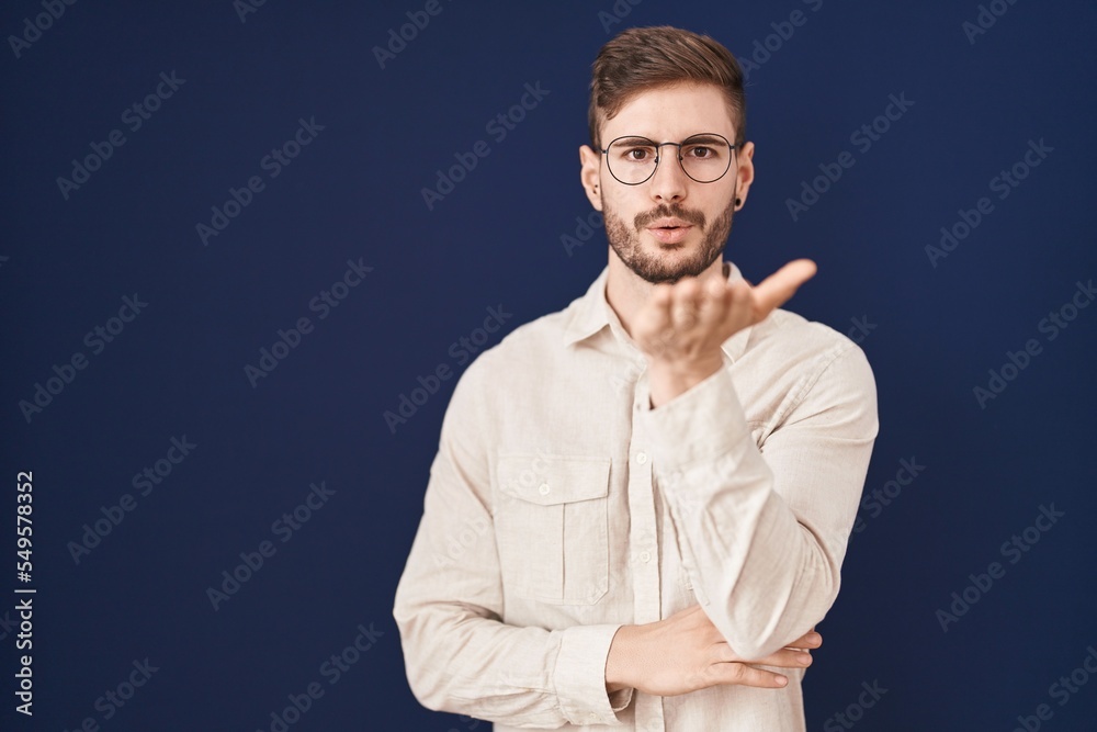 Hispanic man with beard standing over blue background looking at the camera blowing a kiss with hand on air being lovely and sexy. love expression.