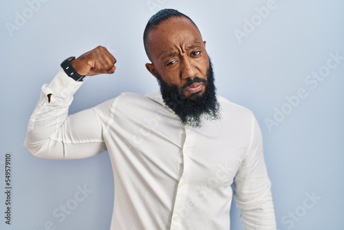African american man standing over blue background strong person showing arm muscle, confident and proud of power