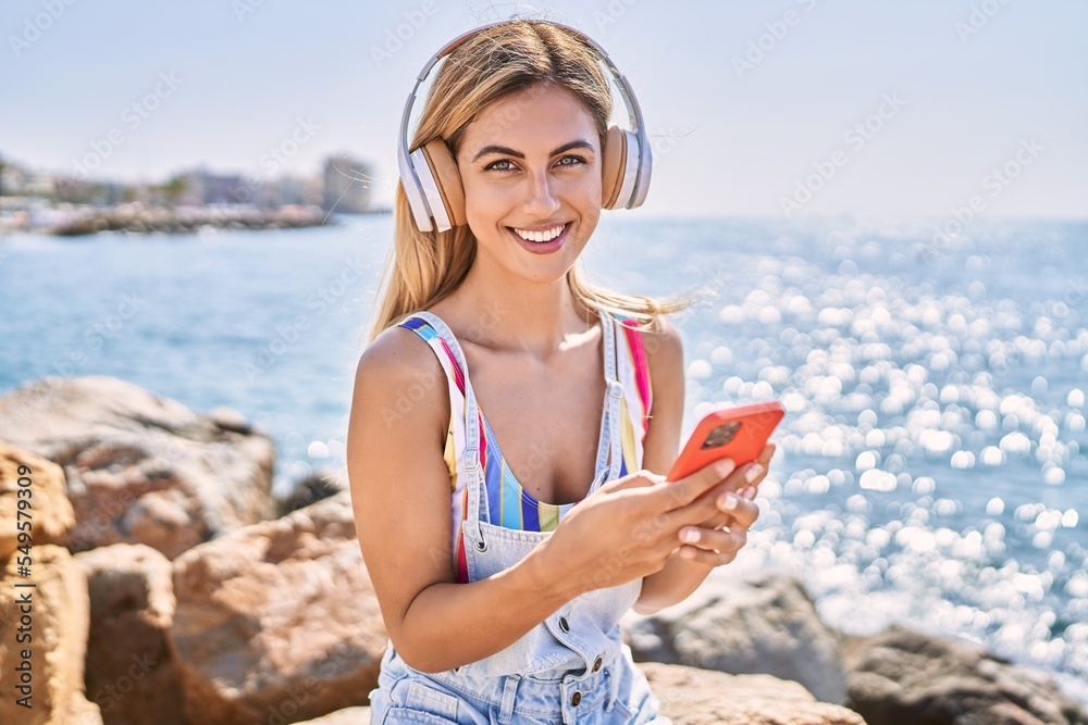 Young blonde girl listening to music sitting on the rock at the beach.