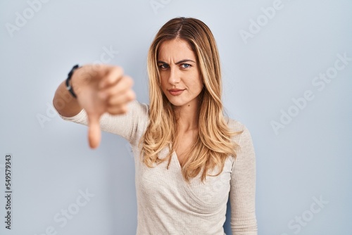 Young blonde woman standing over isolated background looking unhappy and angry showing rejection and negative with thumbs down gesture. bad expression.