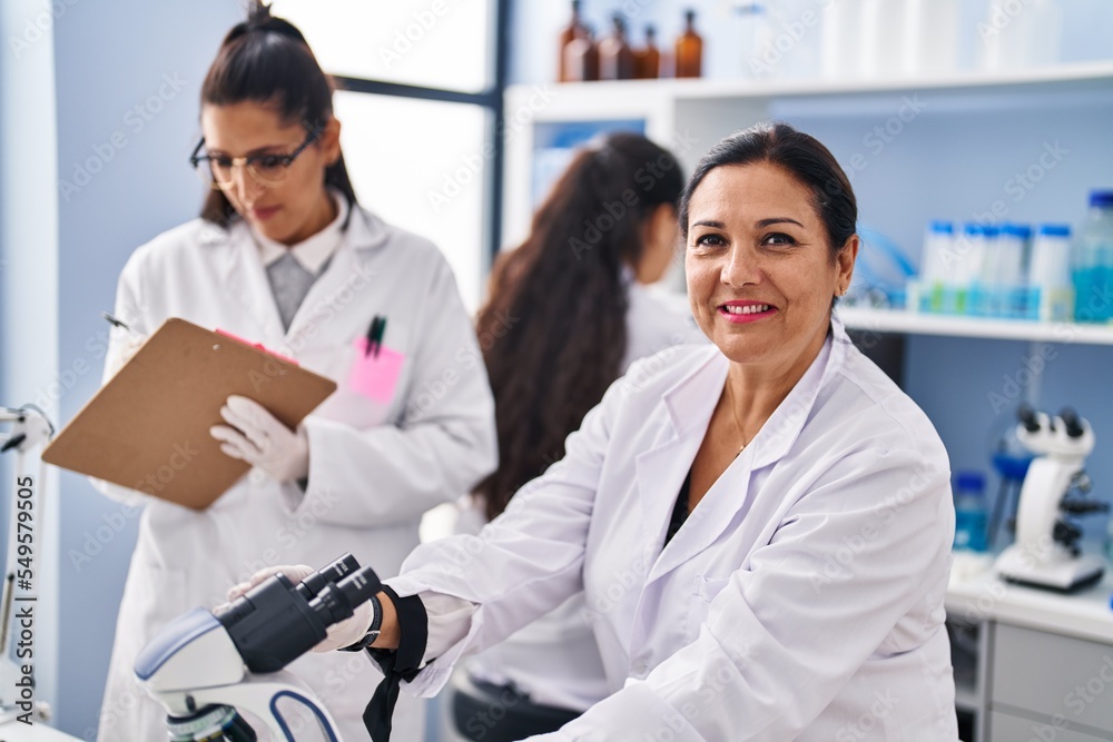 Fototapeta premium Three woman scientists using microscope write on checklist at laboratory